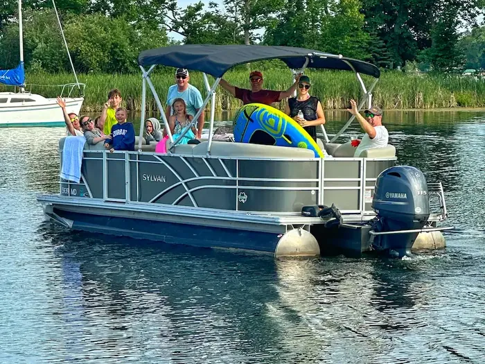 A family waving from the Baxstar Outdoors 22-foot Sylvan pontoon, with a tube aboard and the 115 hp Yamaha outboard